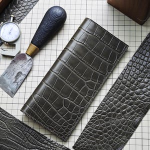 May include: A rectangular olive green leather wallet with a prominent embossed crocodile skin texture. The wallet is positioned on a grid surface alongside a metal leatherworking tool with a dark handle and a small silver dial gauge measuring tool.