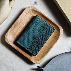 May include: A teal leather wallet with a reptile skin texture rests in a wooden tray. The wallet has a rectangular shape with a textured surface. The tray is made of light-colored wood. The brand name "MESSIEURS" is visible.