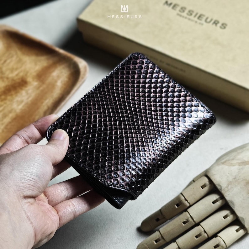May include: A dark brown, textured leather wallet with a scale-like pattern. The wallet is held in a hand, showcasing its design. The brand name "MESSIEURS" is visible on the packaging in the background.