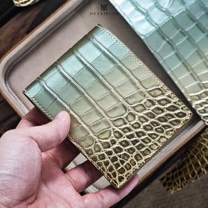 Puede incluir: Cartera de dos pliegues verde y dorada con textura de piel de cocodrilo. La cartera se sostiene en una mano, mostrando el patrón detallado y el degradado de color. La marca "MESSIEURS" es visible en el fondo. Parece ser de cuero.