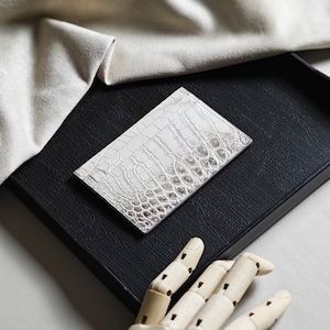 May include: A white cardholder with a crocodile skin effect sits on a black tray. The rectangular cardholder is likely leather. A beige fabric is in the background. The cardholder's dimensions are approximately 10 cm x 7 cm.