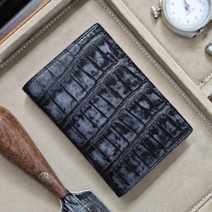 Könnte beinhalten: Eine schwarze Ledergeldbörse mit Krokodilhautstruktur. Die Geldbörse ist rechteckig und geschlossen. Das Bild zeigt auch ein Messgerät und ein Werkzeug mit Holzgriff.