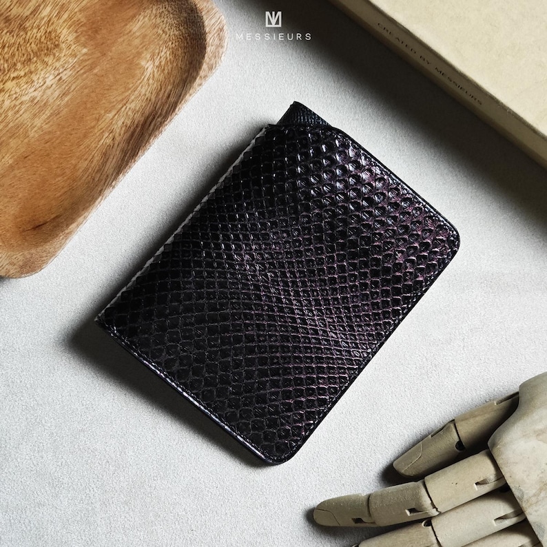 May include: A black, textured wallet with a snakeskin pattern. The wallet is rectangular and appears to be made of leather. The top right corner of the wallet is slightly open. The brand name "MESSIEURS" is printed in white on the top of the wallet.