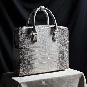 May include: A white crocodile leather briefcase with a top handle and a zip closure. The briefcase has a textured surface and a subtle grey pattern.