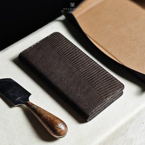 Puede incluir: Una cartera de cuero marrón oscuro con un patrón texturizado y estampado. La cartera está cerrada y se encuentra sobre una superficie blanca.