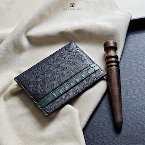 Può includere: Un porta carte in pelle verde scuro con un motivo a struzzo strutturato e una striscia verde in stile coccodrillo. Accanto c'è un utensile in legno. Il marchio "MESSIEURS" è visibile in alto.