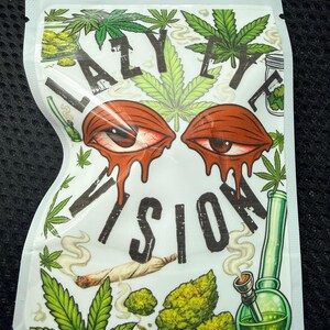 May include: A white resealable pouch featuring the text LAZY EYE VISION in black letters. The design includes two red, bloodshot eyes with dripping effects, green cannabis leaves, a lit joint, and a green glass water pipe with smoke.
