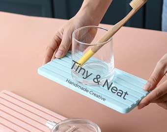 May include: A light blue rectangular tray with parallel grooves, a clear glass tumbler, and a bamboo toothbrush with a yellow handle. The tray features the text Tiny & Neat Handmade Creative. A second pink tray is visible below the blue one.