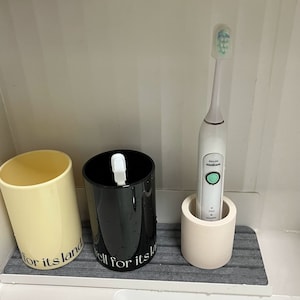 Puede incluir: Dos tazas de cerámica blanca con texto negro, una con el texto "for its land" y la otra con el texto "all for its la", y un cepillo de dientes eléctrico blanco en un soporte de cerámica blanco.