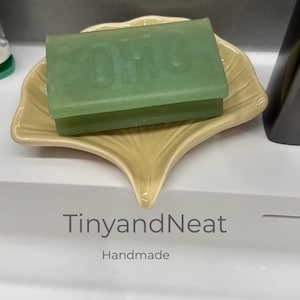 Könnte beinhalten: Eine grüne Seife mit dem aufgedruckten Wort "Olio", die in einer beigefarbenen, muschelförmigen Schale aus Keramik liegt. Die Schale trägt am Boden das Etikett "TinyandNeat Handmade".