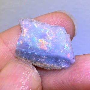 Könnte beinhalten: Ein roher Opal-Edelstein mit einer blau-violetten Grundfarbe, der irisierende Blitze von Grün, Orange und Gelb zeigt. Der Stein hat ein geschichtetes Aussehen und eine natürliche, unpolierte Textur. Der Opal wird in einer Hand gehalten.