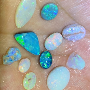 Könnte beinhalten: Eine Sammlung polierter Opal-Edelsteine in verschiedenen Formen und Größen. Die Opale zeigen eine Reihe von Farben, darunter Blau, Grün, Weiß und schillernde Blitze. Einige sind oval, andere unregelmäßig, alle mit einem einzigartigen Farbspiel.