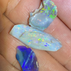 May include: Three raw opal gemstones with irregular shapes. The top stone is light blue with green and orange flashes. The middle stone is elongated, white and blue with rainbow play-of-color. The bottom stone is deep blue with purple tones.