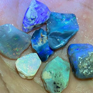 May include: A collection of raw opal gemstones in various shades of blue, green, and white. The opals have a rough, natural texture and display iridescent flashes of color. The gemstones are held in a hand, showcasing their unique shapes and colors.