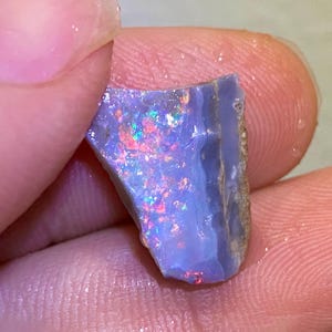 Könnte beinhalten: Ein roher Opal-Edelstein mit einer blau-grauen Grundfarbe. Der Opal zeigt lebhafte Blitze in Rot, Grün und Gelb, wodurch ein Farbspiel-Effekt entsteht. Der Stein wird zwischen Fingern gehalten und zeigt seinen natürlichen, unpolierten Zustand.