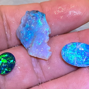 May include: Three opals are shown, each with unique shapes and colours. A rough opal is a pale blue with green and blue flashes. An oval opal is a deep green with bright green and blue flashes. Another oval opal is a bright blue with green flashes.