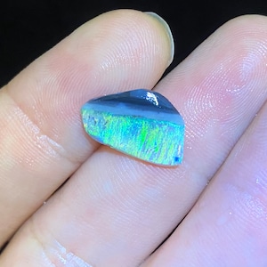 May include: A small, iridescent opal gemstone with a black and white base and a green and blue flash. The opal is set in a clear resin.
