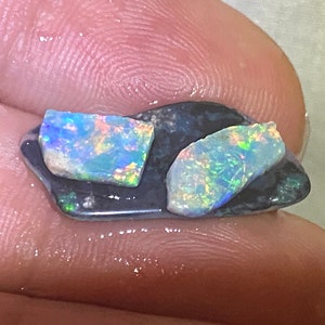 May include: Two iridescent opal gemstones mounted on a black backing. The opals are a light blue and green colour with flashes of rainbow.