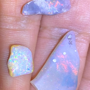 May include: Three iridescent opal pieces with a play of color. The opals display a range of colors, including blue, pink, and yellow, with a shimmering effect. The shapes are irregular, showcasing the natural beauty of the gemstones.