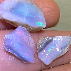 May include: Three raw opal gemstones with iridescent flashes of color. The opals display a range of colors, including blue, green, and pink, against a milky white and tan background. The rough texture of the stones is visible.