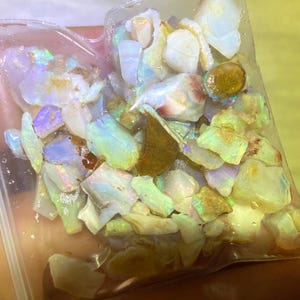 May include: A plastic bag filled with a variety of small, iridescent opal stones. The stones are a mix of white, green, and blue.
