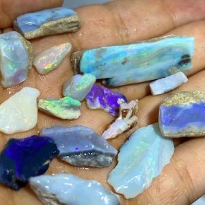 Könnte beinhalten: Eine Sammlung roher Opal-Edelsteine in verschiedenen Formen und Größen. Die Opale zeigen eine Reihe von Farben, darunter Blau, Weiß, Lila und Grün, mit schillernden Blitzen. Einige Opale haben eine raue, natürliche Textur, während andere glatter erscheinen.