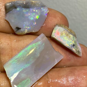 Könnte beinhalten: Drei Stücke roher Opal, die eine Reihe von Farben zeigen, darunter Blau, Grün und Blitze von schillerndem Licht. Die Opalstücke haben eine raue, natürliche Textur und unterschiedliche Formen. Der Hintergrund ist neutral, wodurch die Edelsteine hervorgehoben werden.