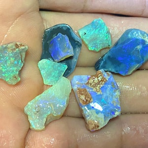 May include: A collection of seven rough opal gemstones in various shades of blue, green, and white. The opals are iridescent and have a natural, unpolished finish.
