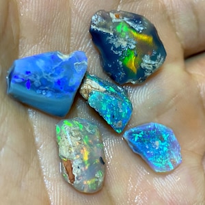 Könnte beinhalten: Eine Sammlung roher Opal-Edelsteine in verschiedenen Blau-, Grün- und Gelbtönen. Die Opale haben eine raue, natürliche Textur und zeigen schillernde Farbblitze. Die Edelsteine werden in einer Hand gehalten, um ihre einzigartigen Muster und Farbtöne zu präsentieren.