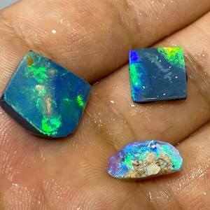 Könnte beinhalten: Drei rohe Opal-Edelsteine mit lebendigen blauen, grünen und violetten Blitzen. Die Opale werden auf einer hellen Oberfläche präsentiert, die ihre natürlichen, unregelmäßigen Formen und einzigartigen Farbmuster hervorhebt. Diese Edelsteine sind ideal für die Schmuckherstellung.