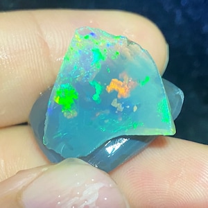 Könnte beinhalten: Ein hellblauer Opal-Edelstein mit grünen und orangefarbenen Farbschimmern. Der Opal ist in einer dunkelgrauen Basis gefasst.