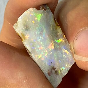 Könnte beinhalten: Ein roher Opal-Edelstein mit weißer Grundfarbe und schillernden Blitzen in Grün, Gelb und Blau. Der Naturstein hat eine strukturierte Oberfläche und wird zwischen Fingern gehalten, was sein einzigartiges Farbspiel zeigt.