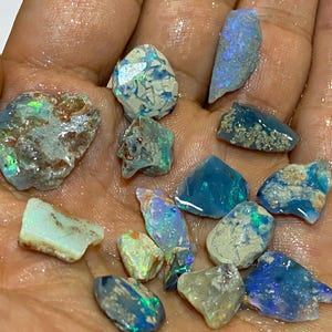 Op de afbeelding: Een verzameling ruwe opaal edelstenen in verschillende vormen en maten. De opalen vertonen een scala aan kleuren, waaronder blauw, groen en flitsen van iriserend licht. Sommige opalen hebben een melkachtige witte basis met blauwe en groene patronen.