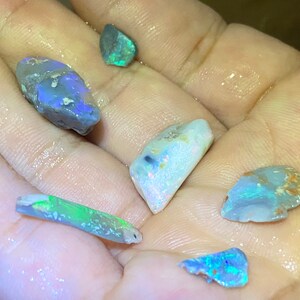 May include: A collection of five rough opal stones in various shades of blue, green, and white. The stones are iridescent and have a natural, unpolished finish.