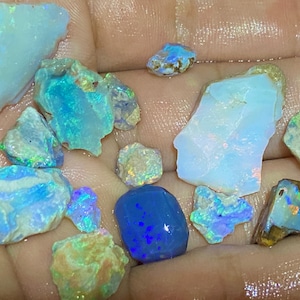 Könnte beinhalten: Eine Sammlung roher Opale in verschiedenen Formen und Größen, die ein Farbspektrum von Blau, Grün und schillernden Lichtblitzen zeigen. Die Opale werden in einer Hand gehalten, was ihre natürliche Schönheit hervorhebt.