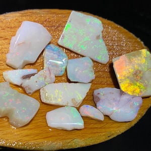 May include: A collection of irregular, white-toned opal gemstones with iridescent flashes of green, orange, and pink, resting on a smooth, oval-shaped wooden surface. The stones vary in size and shape, each displaying unique light-reflecting properties.