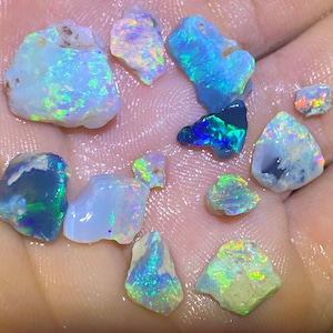 Könnte beinhalten: Eine Sammlung kleiner, irisierender Opal-Edelsteine in verschiedenen Blautönen, Grüntönen und Weiß. Die Opale sind roh und haben eine natürliche, unpolierte Oberfläche.