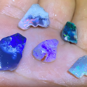 May include: Five rough-cut opal gemstones in various shades of blue, purple, and white. The opals are displayed on a hand.