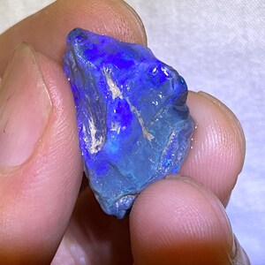 Könnte beinhalten: Ein roher, blaugrüner Opal mit irisierenden Blitzen von Lila und Blau. Der Opal ist ein natürlicher Edelstein und hat eine einzigartige, unregelmäßige Form.