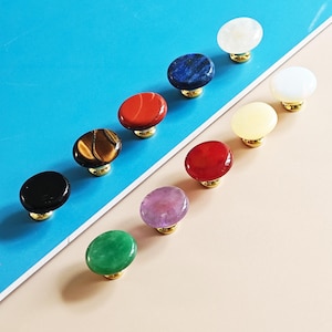 May include: A collection of oval-shaped cabinet knobs in various colors and textures. The knobs feature a polished stone appearance, including black, tiger's eye, red, blue, and green. Each knob is mounted on a gold-colored base.