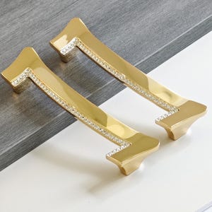 Könnte beinhalten: Zwei goldfarbene Schrankgriffe mit rechteckiger Form und einer dekorativen Reihe kleiner, klarer Kristalle. Die Griffe haben eine glänzende Oberfläche und sind vor einem neutralen Hintergrund platziert.