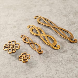 May include: An assortment of antique brass cabinet hardware, including two styles of pulls and two styles of knobs. The pulls feature an infinity symbol design, while the knobs have a floral-like pattern. The hardware is displayed on a gray surface.