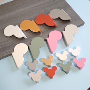 Puede incluir: Una colección de coloridos tiradores y pomos de madera para armarios en varias formas. Los pomos tienen forma de cabeza de ratón y los tiradores tienen una forma única. Los colores incluyen blanco, gris, naranja, rosa y verde.