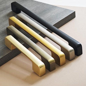 Pode incluir: Uma coleção de puxadores de armário em vários acabamentos metálicos, incluindo dourado, bronze, prateado e preto. Os puxadores estão dispostos diagonalmente, mostrando suas diferentes cores e designs. O hardware é exibido em um fundo neutro.