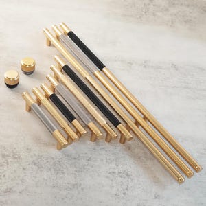 May include: A selection of gold-coloured cabinet pulls and knobs. The pulls have textured handles in gold, silver, and black, with cylindrical and bar-shaped designs. The knobs are small, round, and gold. The hardware is arranged on a light grey surface.