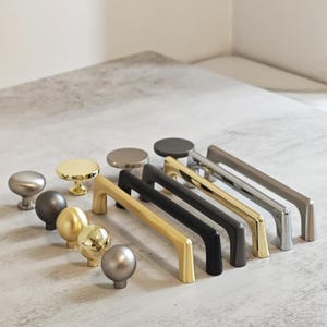May include: A selection of cabinet hardware, including knobs and pulls, in various metallic finishes. The selection features round knobs in silver, gold, and dark grey, along with bar pulls in gold, black, and silver. The hardware is designed for furniture and cabinetry.