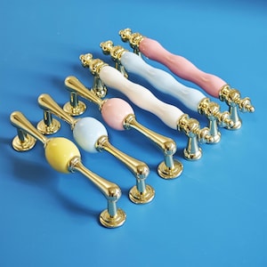 Op de afbeelding: Een verzameling kleurrijke kastgrepen met goudkleurige hardware. De grepen hebben een verscheidenheid aan pastelkleuren, waaronder roze, blauw, geel en wit. De handgrepen hebben een decoratief ontwerp.