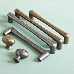 May include: An assortment of cabinet hardware, including two oval knobs and four bar pulls. The knobs and pulls have a metallic finish in shades of bronze and silver. The hardware is arranged on a light green surface.