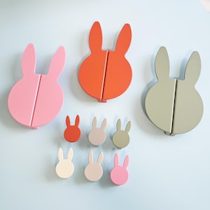 Op de afbeelding: Een verzameling konijnvormige wandhaken in verschillende maten en kleuren, waaronder roze, oranje en groen. Ze zijn bevestigd aan een lichtblauwe muur. Het ontwerp is modern en speels, ideaal voor een kinderkamer of een speelse ruimte.