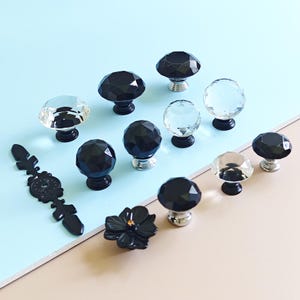 Op de afbeelding: Een verzameling decoratieve kastknoppen en -grepen in verschillende stijlen. De knoppen hebben facetgeslepen ontwerpen van zwart en helder kristal, met zilveren en zwarte bases. Een zwarte bloemenknop en een decoratieve trekker zijn ook inbegrepen.